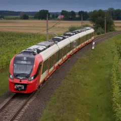 Czerwony pociąg SKM jedzie po torach między polami kukurydzy i zboża, w tle domy i las. Trasa Warszawa-Legionowo.