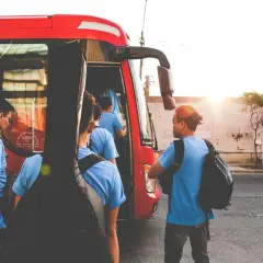 Grupa młodych ludzi z gitarą wsiada do czerwonego autobusu GTV. Słońce zachodzi, tworząc ciepłą atmosferę.