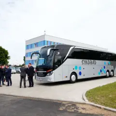 Grupa mężczyzn w garniturach rozmawia przy autokarze Sindbad. Sprawdzają ceny biletów przed podróżą.