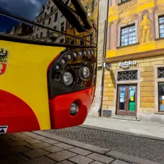 Autobus kraków komunikacja miejska z herbem miasta. Na żółto-czerwonym pojeździe widoczny herb Krakowa. W tle kamienica z malowidłami i sklep Żabka.