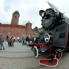 Czarna lokomotywa TLK Pirat przy ceglanej twierdzy z latarnią morską. Tłum ludzi ogląda parowóz.