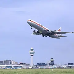 Samolot McDonnell Douglas DC-10 wznosi się nad lotniskiem Schiphol. W tle widoczna wieża kontroli lotów i inne samoloty.