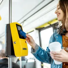 Uśmiechnięta kobieta z kawą w ręku zbliża kartę do żółtego kasownika, aby aktywować swój bilet okresowy w autobusie.
