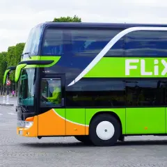 Zielony autokar Flixbus z pomarańczowym pasem na dole, jadący ulicą. Kierowca w żółtej kurtce. Czy Flixbus robi postoje? Tak, ten autokar jest w ruchu.