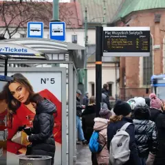 Kraków, Plac Wszystkich Świętych. Ludzie czekają na tramwaj, obok automat z biletami. W tle zabytkowa architektura i niebieski tramwaj.