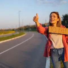 Młoda kobieta z uśmiechem na twarzy łapie autostop. Trzyma tabliczkę "TO FREEDOM" i kciuk w górę, gotowa na przygodę.