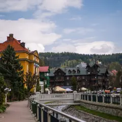 Urokliwa promenada w Krynicy-Zdroju, z widokiem na zabytkowe wille i górskie zbocza. W oddali widać budynek z napisem "Polonus", przypominający o podróży z Warszawy Zachodniej.