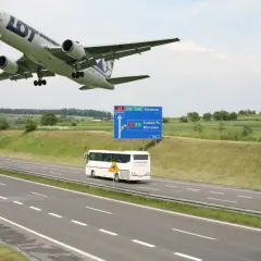 Samolot LOT leci nisko nad autostradą A4 w kierunku Katowic. Obok jedzie autobus, a w oddali widać drogę do Krakowa i Warszawy.