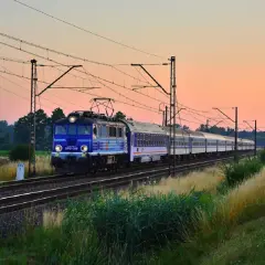 Niebieski pociąg PKP EP07-389 z wagonami mija pole o zachodzie słońca, jadąc w stronę Kołobrzegu.