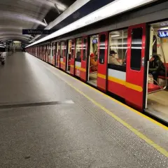 Czerwony pociąg metra M1 z otwartymi drzwiami na stacji. Pasażerowie czekają na peronie.