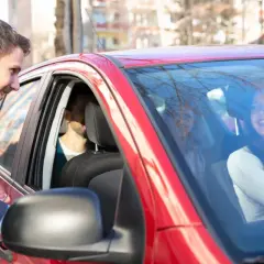 Mężczyzna w garniturze pochyla się do czerwonego samochodu, by porozmawiać z uśmiechniętą kobietą za kierownicą. To świetny przykład carpoolingu!