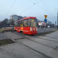 Czerwony ztm 26 tramwaj przejeżdża przez skrzyżowanie. W tle widać autobus i budynki.