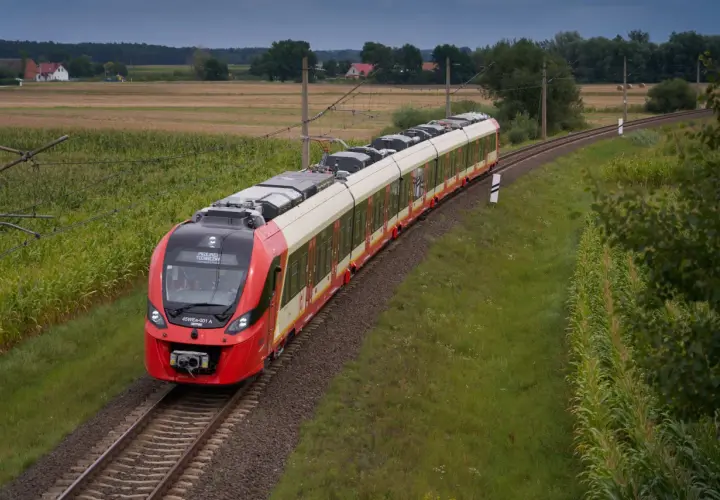Czerwony pociąg SKM jedzie po torach między polami kukurydzy i zboża, w tle domy i las. Trasa Warszawa-Legionowo.