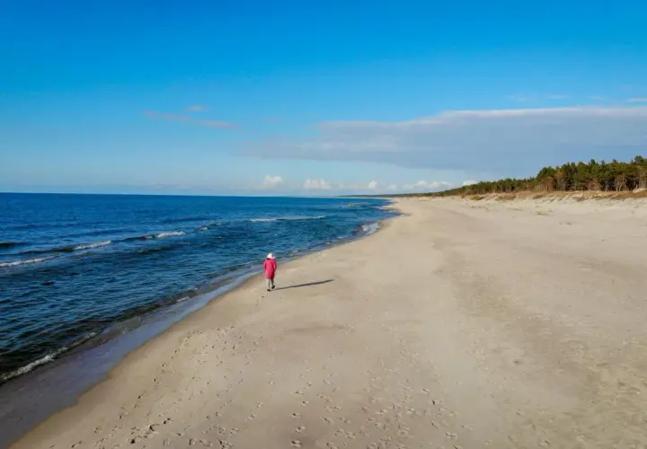 Samotna postać w różowym płaszczu spaceruje po plaży w Krynicy Morskiej. Wyobraź sobie, że to ostatni wagon pociągu do raju.