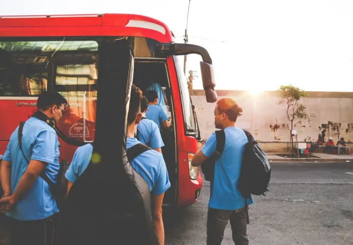 Grupa młodych ludzi z gitarą wsiada do czerwonego autobusu GTV. Słońce zachodzi, tworząc ciepłą atmosferę.