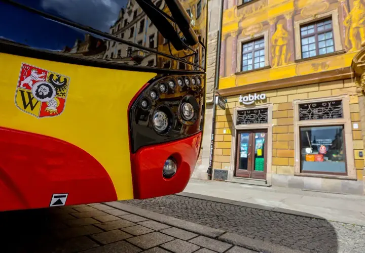 Autobus kraków komunikacja miejska z herbem miasta. Na żółto-czerwonym pojeździe widoczny herb Krakowa. W tle kamienica z malowidłami i sklep Żabka.