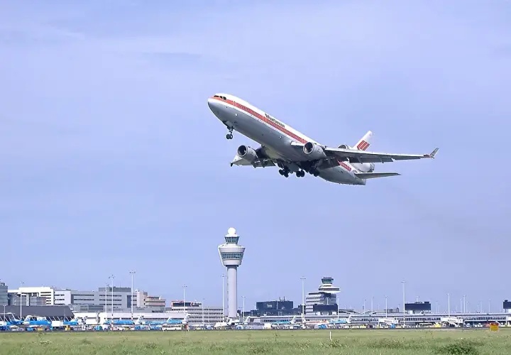 Samolot McDonnell Douglas DC-10 wznosi się nad lotniskiem Schiphol. W tle widoczna wieża kontroli lotów i inne samoloty.