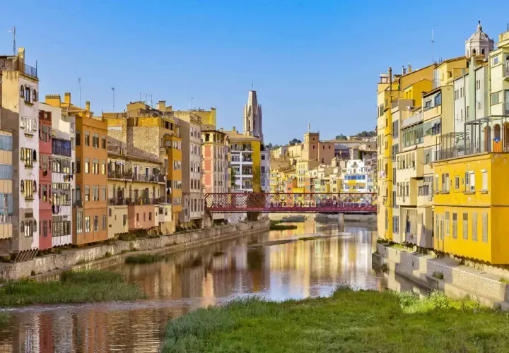 Kolorowe kamienice nad rzeką w Gironie, z mostem łączącym z Barceloną w tle.