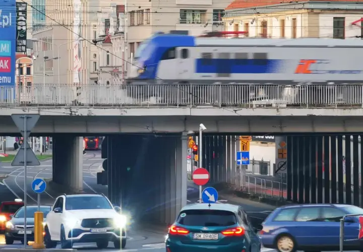 Pędzący pociąg IC 407 na trasie Chopin mija ruchliwe skrzyżowanie z samochodami i tramwajami w tle.