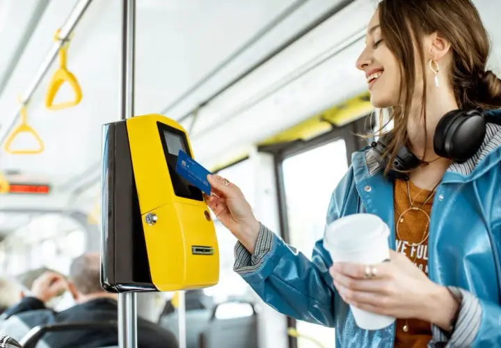 Uśmiechnięta kobieta z kawą w ręku zbliża kartę do żółtego kasownika, aby aktywować swój bilet okresowy w autobusie.