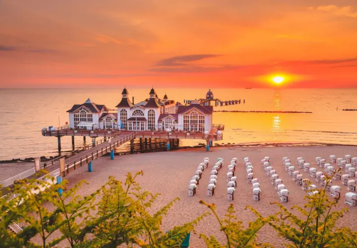 Zachód słońca nad Rugią, idealny cel podróży z Polski. Molo z białymi budynkami, plaża z rzędami koszy plażowych.