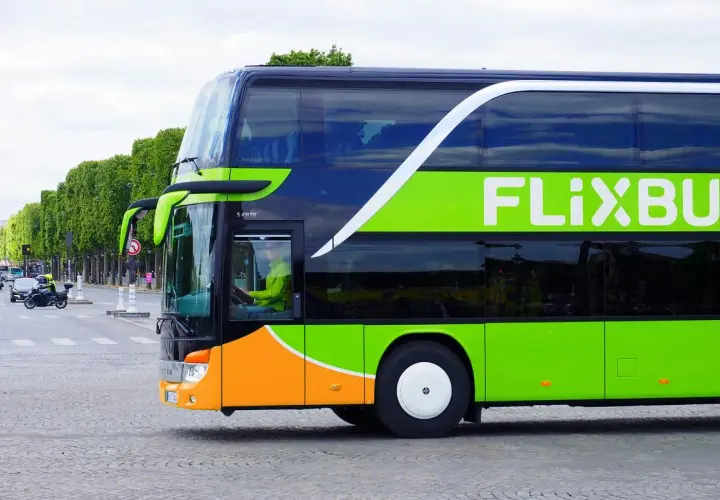 Zielony autokar Flixbus z pomarańczowym pasem na dole, jadący ulicą. Kierowca w żółtej kurtce. Czy Flixbus robi postoje? Tak, ten autokar jest w ruchu.