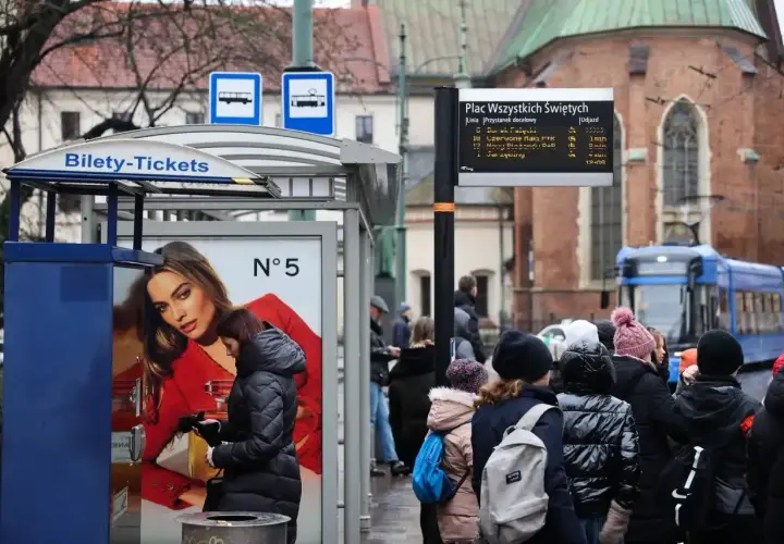 Kraków, Plac Wszystkich Świętych. Ludzie czekają na tramwaj, obok automat z biletami. W tle zabytkowa architektura i niebieski tramwaj.