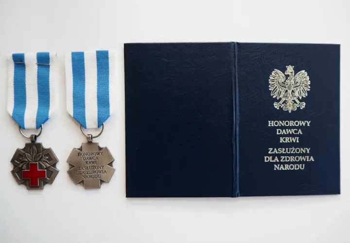 Dwa medale z czerwonym krzyżem i napisem "Honorowy Dawca Krwi Zasłużony dla Zdrowia Narodu" oraz granatowa teczka z godłem Polski.