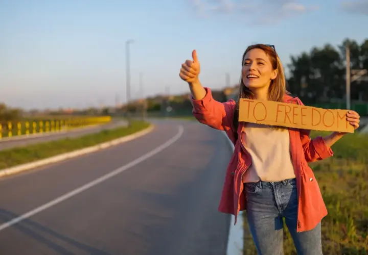 Młoda kobieta z uśmiechem na twarzy łapie autostop. Trzyma tabliczkę "TO FREEDOM" i kciuk w górę, gotowa na przygodę.