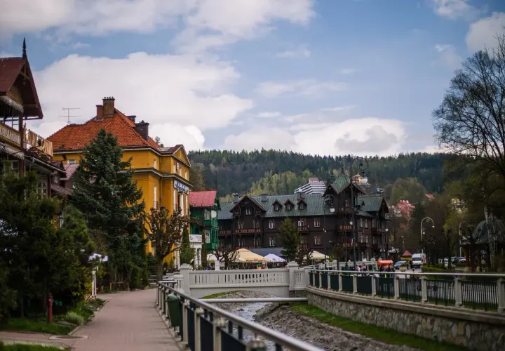 Urokliwa promenada w Krynicy-Zdroju, z widokiem na zabytkowe wille i górskie zbocza. W oddali widać budynek z napisem "Polonus", przypominający o podróży z Warszawy Zachodniej.