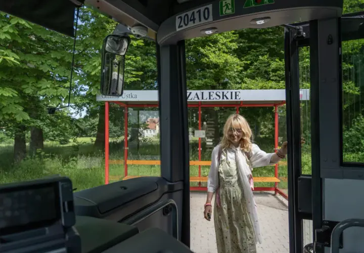 Młoda kobieta wsiada do autobusu na przystanku w gminie Ustka. W tle zielony krajobraz i budynek.