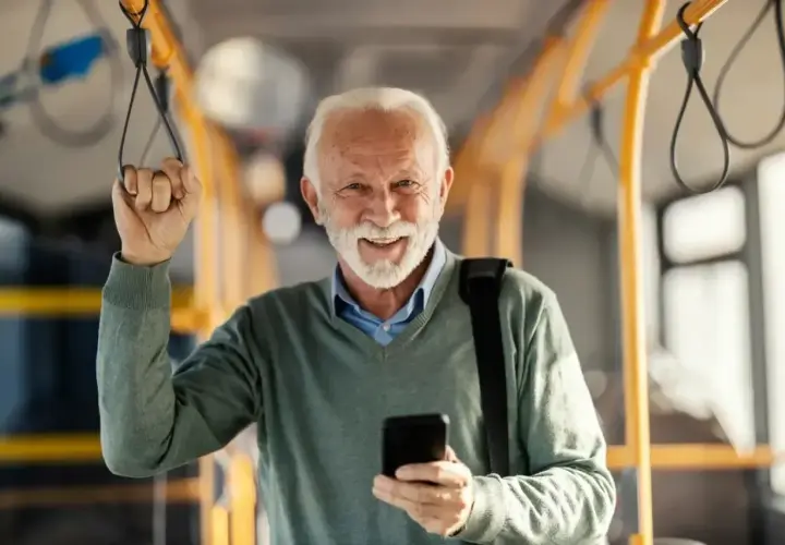 Uśmiechnięty starszy pan z telefonem w dłoni, trzymający się poręczy w pojeździe. Cieszy się z możliwości 70 lat darmowe przejazdy PKP Intercity.