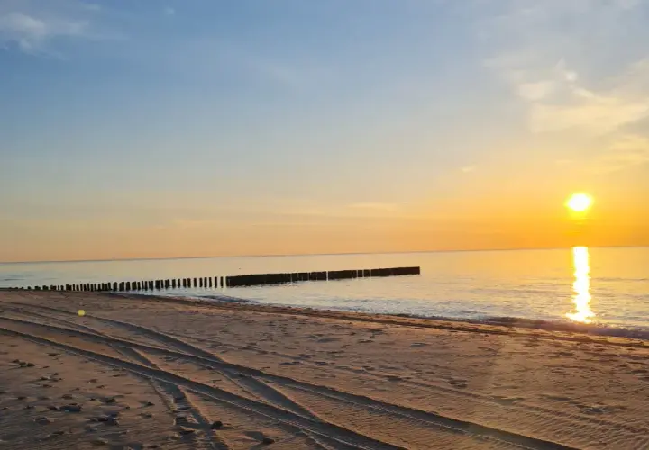 Zachód słońca nad morzem, piaszczysta plaża z widocznymi śladami opon, falochrony. Idealne miejsce na odpoczynek po podróży, nawet jeśli wicie dojazd PKP.