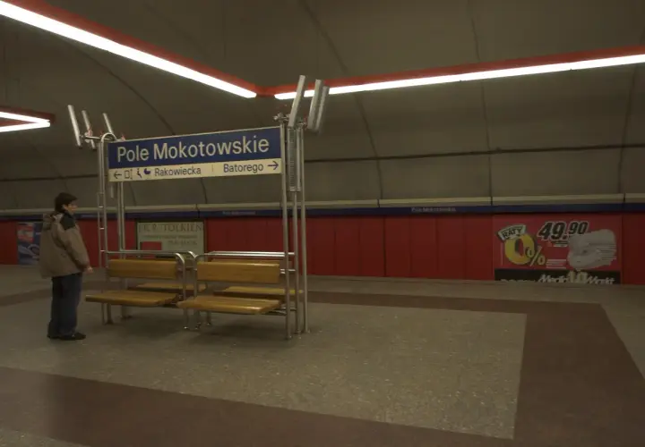 Na stacji metra Pole Mokotowskie stoi mężczyzna obok ławki. W tle niebieska tablica z nazwami ulic i czerwone panele.