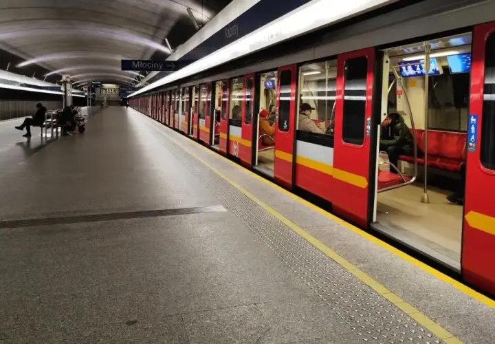 Czerwony pociąg metra M1 z otwartymi drzwiami na stacji. Pasażerowie czekają na peronie.