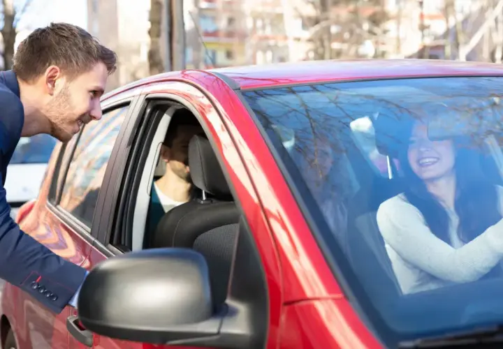 Mężczyzna w garniturze pochyla się do czerwonego samochodu, by porozmawiać z uśmiechniętą kobietą za kierownicą. To świetny przykład carpoolingu!
