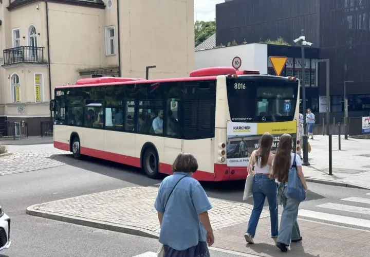 Autobus linii 148 w Gdańsku, z numerem bocznym 8016, odjeżdża z przystanku. Na pierwszym planie dwie dziewczyny idące przez pasy.