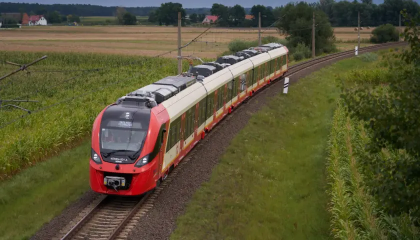 Czerwony pociąg SKM jedzie po torach między polami kukurydzy i zboża, w tle domy i las. Trasa Warszawa-Legionowo.