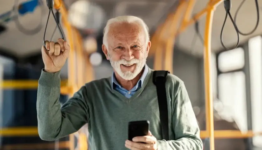 Uśmiechnięty starszy pan w autobusie, korzystający z ulgi dla emerytów w Wałbrzychu. Trzyma telefon i poręcz.