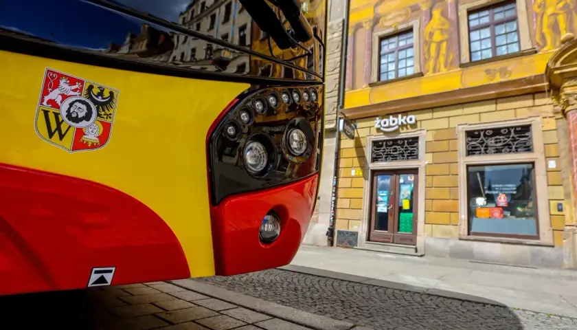 Autobus kraków komunikacja miejska z herbem miasta. Na żółto-czerwonym pojeździe widoczny herb Krakowa. W tle kamienica z malowidłami i sklep Żabka.