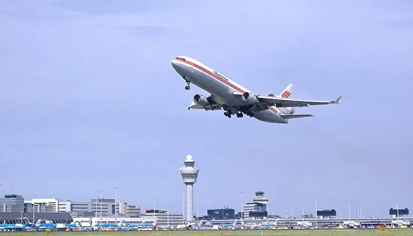 Samolot McDonnell Douglas DC-10 wznosi się nad lotniskiem Schiphol. W tle widoczna wieża kontroli lotów i inne samoloty.