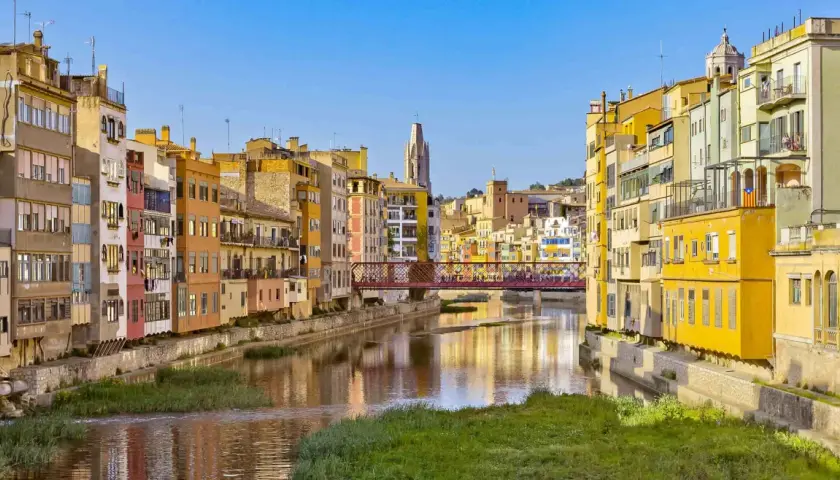Kolorowe kamienice nad rzeką w Gironie, z mostem łączącym z Barceloną w tle.