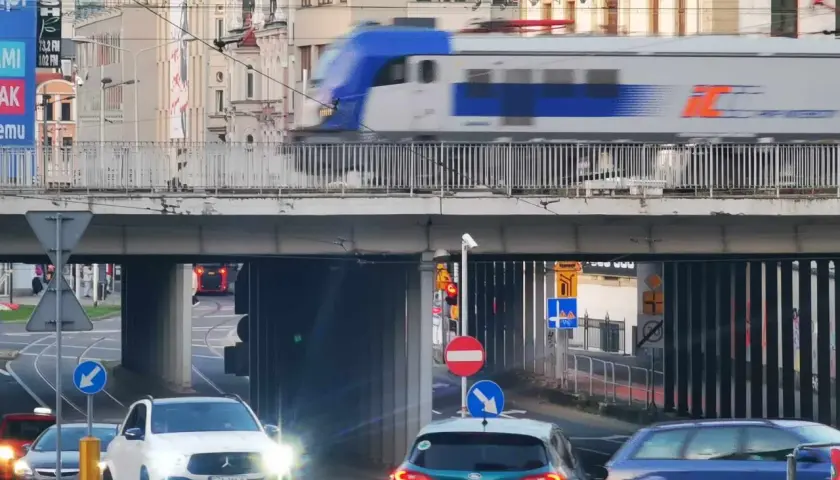 Pędzący pociąg IC 407 na trasie Chopin mija ruchliwe skrzyżowanie z samochodami i tramwajami w tle.