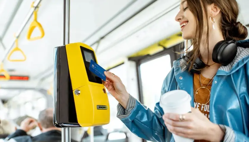 Uśmiechnięta kobieta z kawą w ręku zbliża kartę do żółtego kasownika, aby aktywować swój bilet okresowy w autobusie.