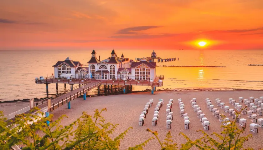 Zachód słońca nad Rugią, idealny cel podróży z Polski. Molo z białymi budynkami, plaża z rzędami koszy plażowych.