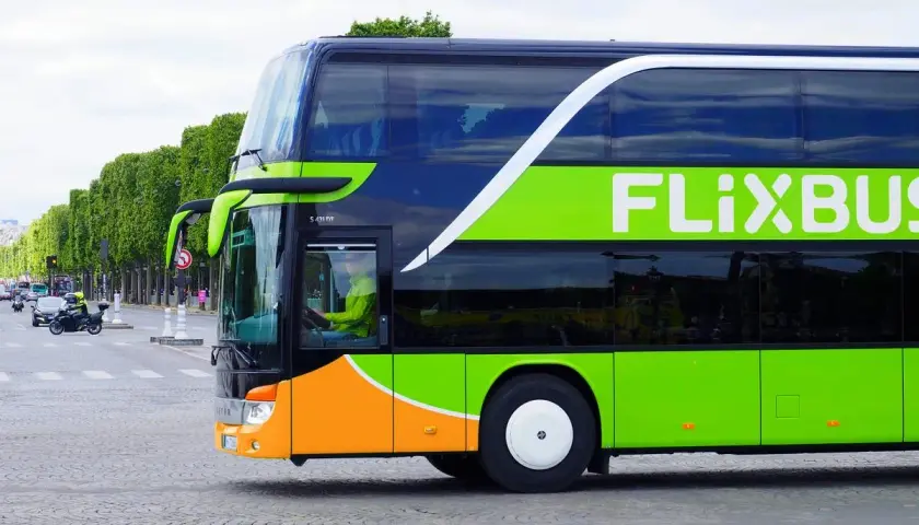 Zielony autokar Flixbus z pomarańczowym pasem na dole, jadący ulicą. Kierowca w żółtej kurtce. Czy Flixbus robi postoje? Tak, ten autokar jest w ruchu.
