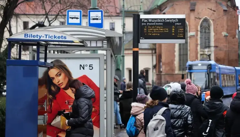 Kraków, Plac Wszystkich Świętych. Ludzie czekają na tramwaj, obok automat z biletami. W tle zabytkowa architektura i niebieski tramwaj.