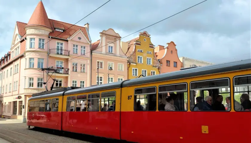 Czerwono-żółty tramwaj z numerem 87 jedzie ulicą Grudziądza. W tle kolorowe kamienice.