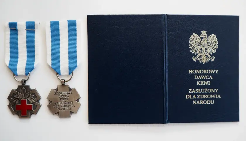 Dwa medale z czerwonym krzyżem i napisem "Honorowy Dawca Krwi Zasłużony dla Zdrowia Narodu" oraz granatowa teczka z godłem Polski.