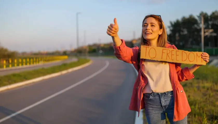 Młoda kobieta z uśmiechem na twarzy łapie autostop. Trzyma tabliczkę "TO FREEDOM" i kciuk w górę, gotowa na przygodę.